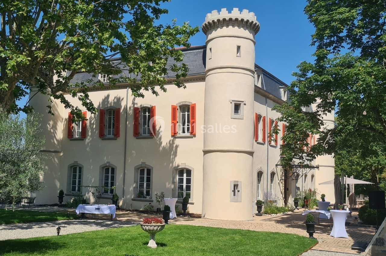Façade d'un château avec tour ronde, volets rouges, entouré d'arbres et d'une pelouse bien entretenue.