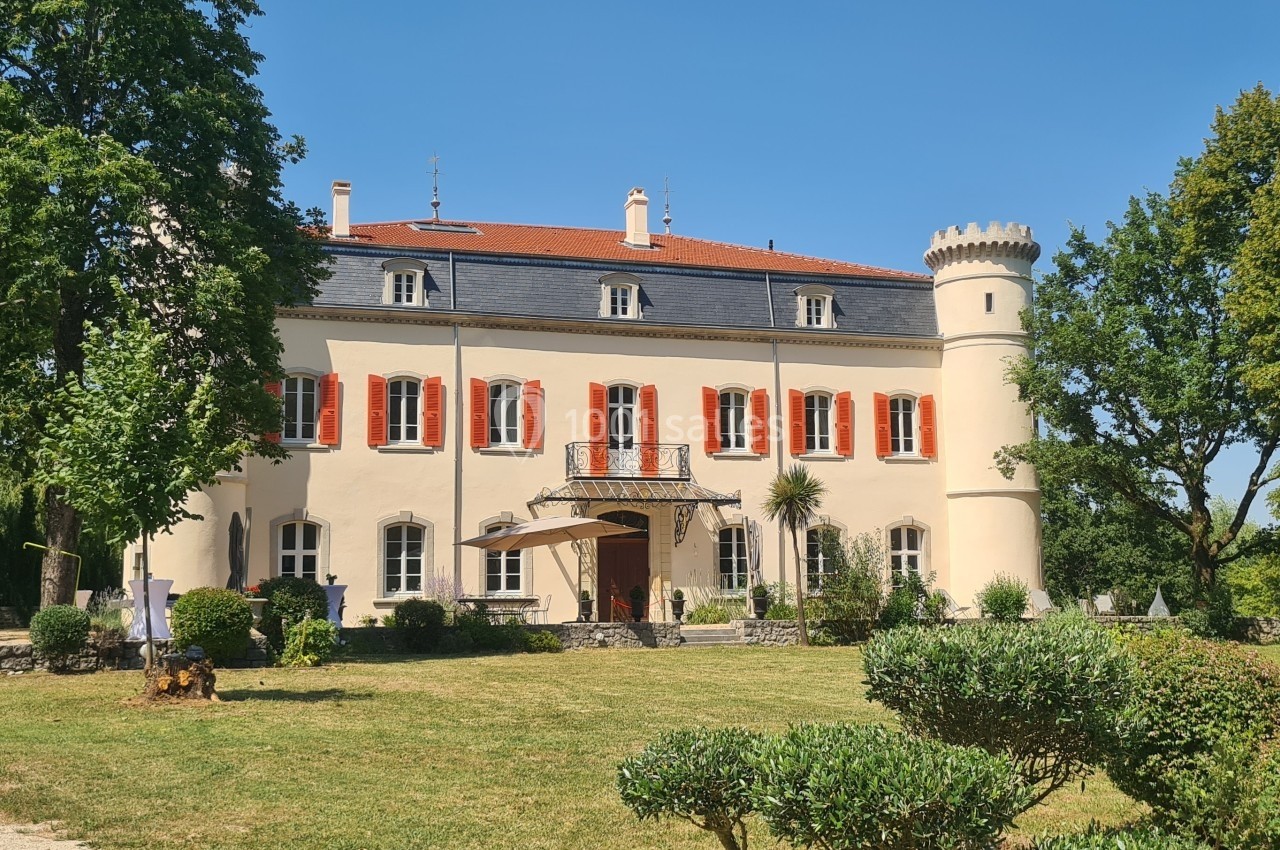 Façade d'un château avec tour ronde, entouré d'un jardin arboré sous un ciel bleu.