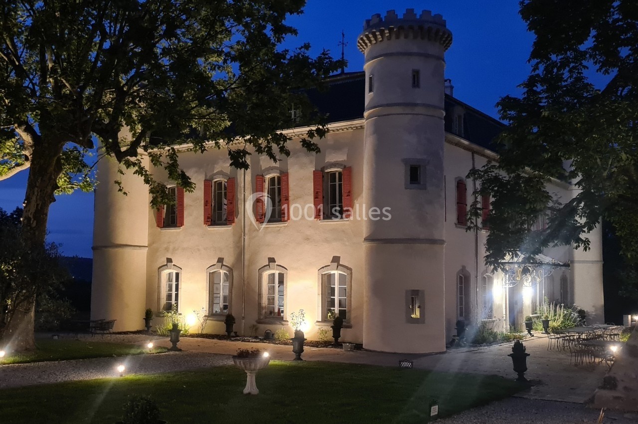 Façade d'un château éclairé en soirée, entouré d'arbres et d'un jardin avec éclairage au sol.
