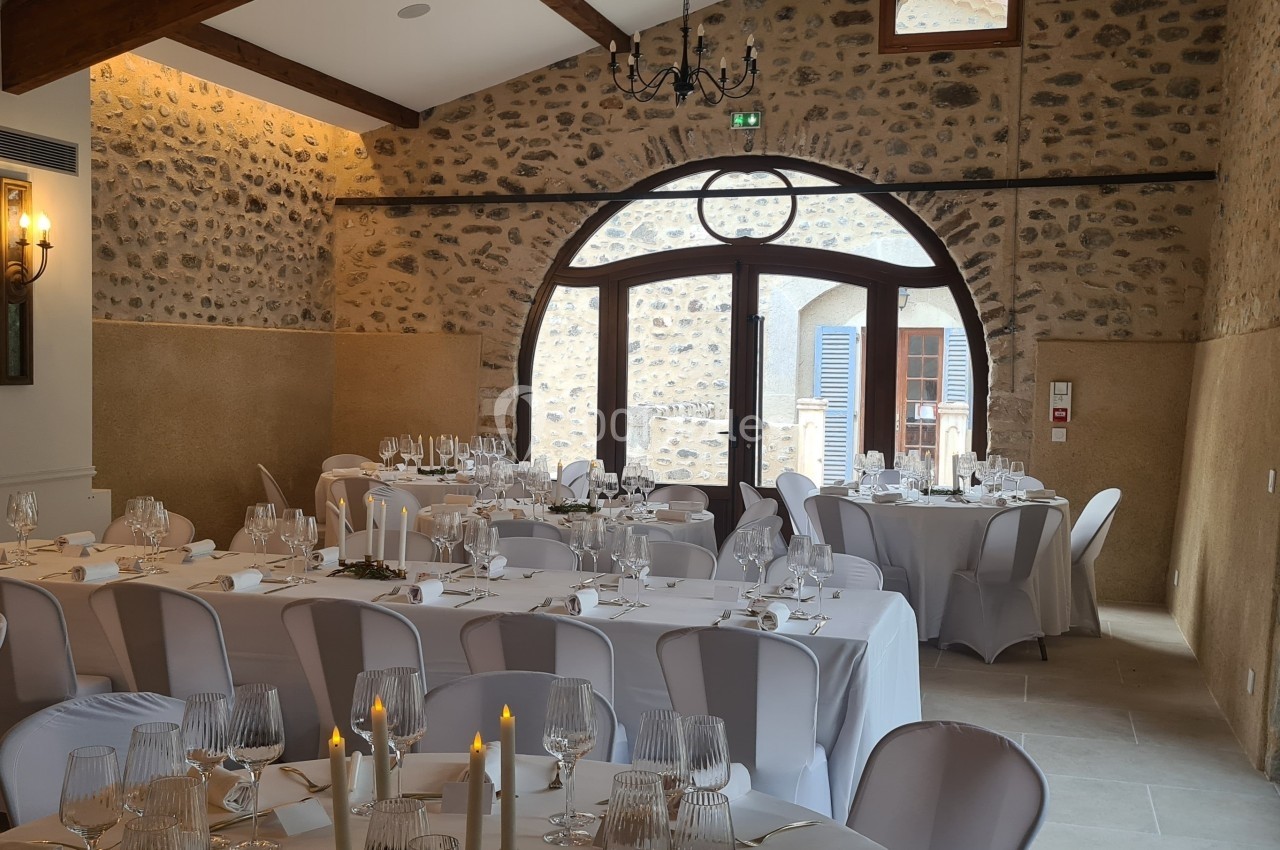 Salle de réception en pierre avec tables dressées, nappes blanches et chandeliers, éclairée par des fenêtres et une porte…
