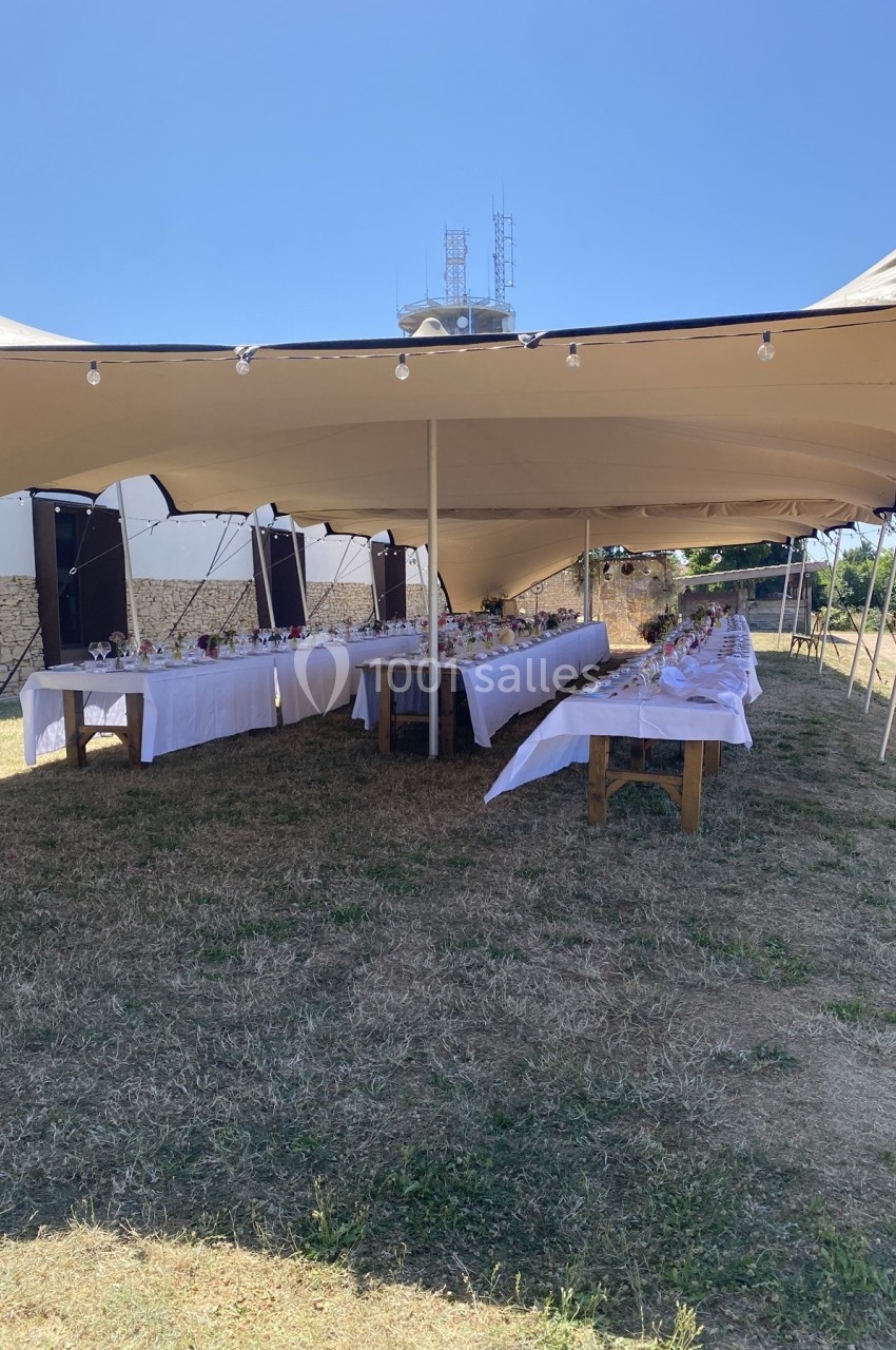 Tables dressées sous une grande tente beige en extérieur, avec nappes blanches et vaisselle disposée pour un événement.