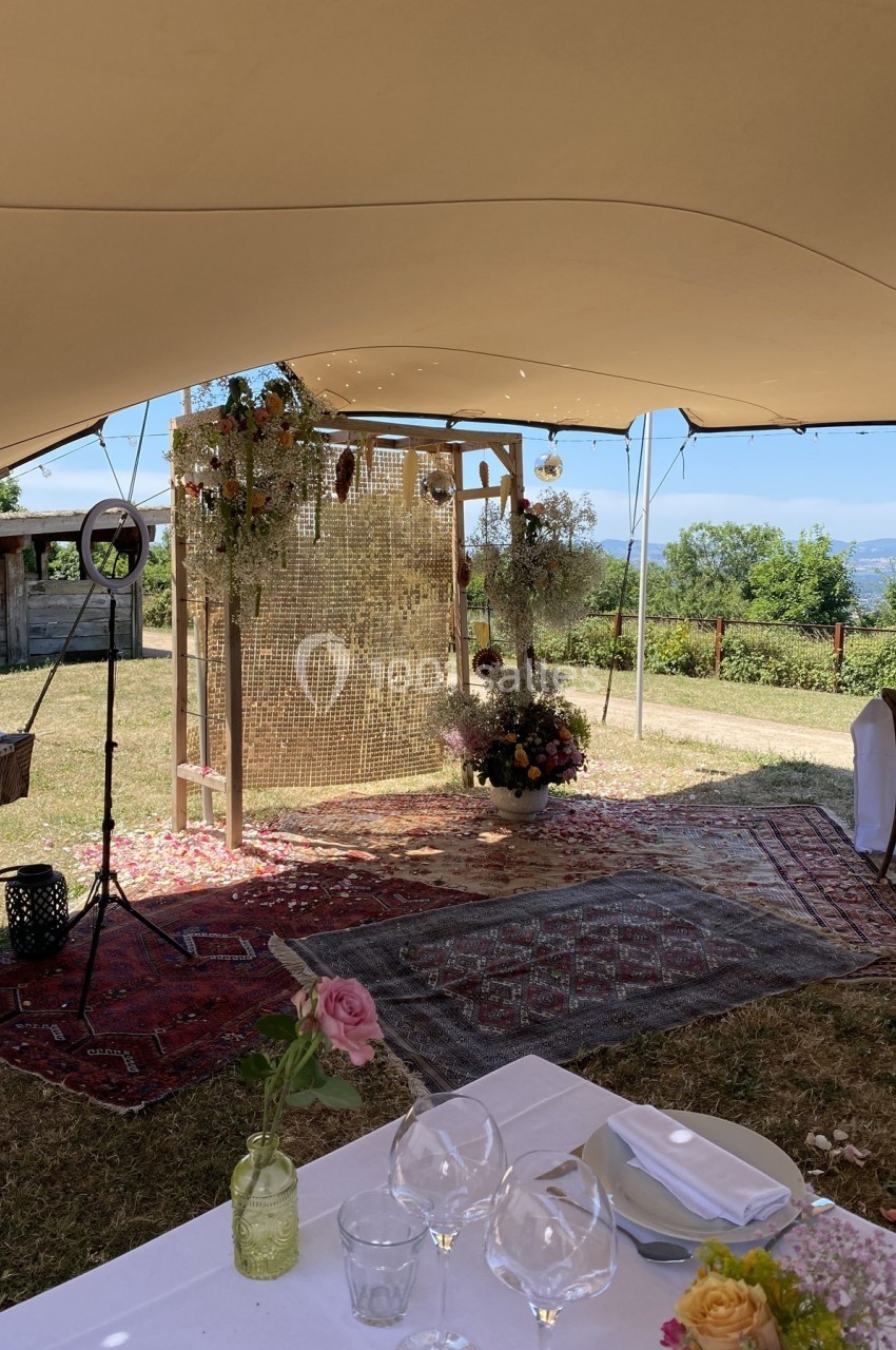 Décor champêtre avec arche ornée de fleurs et rideau doré, sous une tente, entouré de tapis et de pétales au sol.