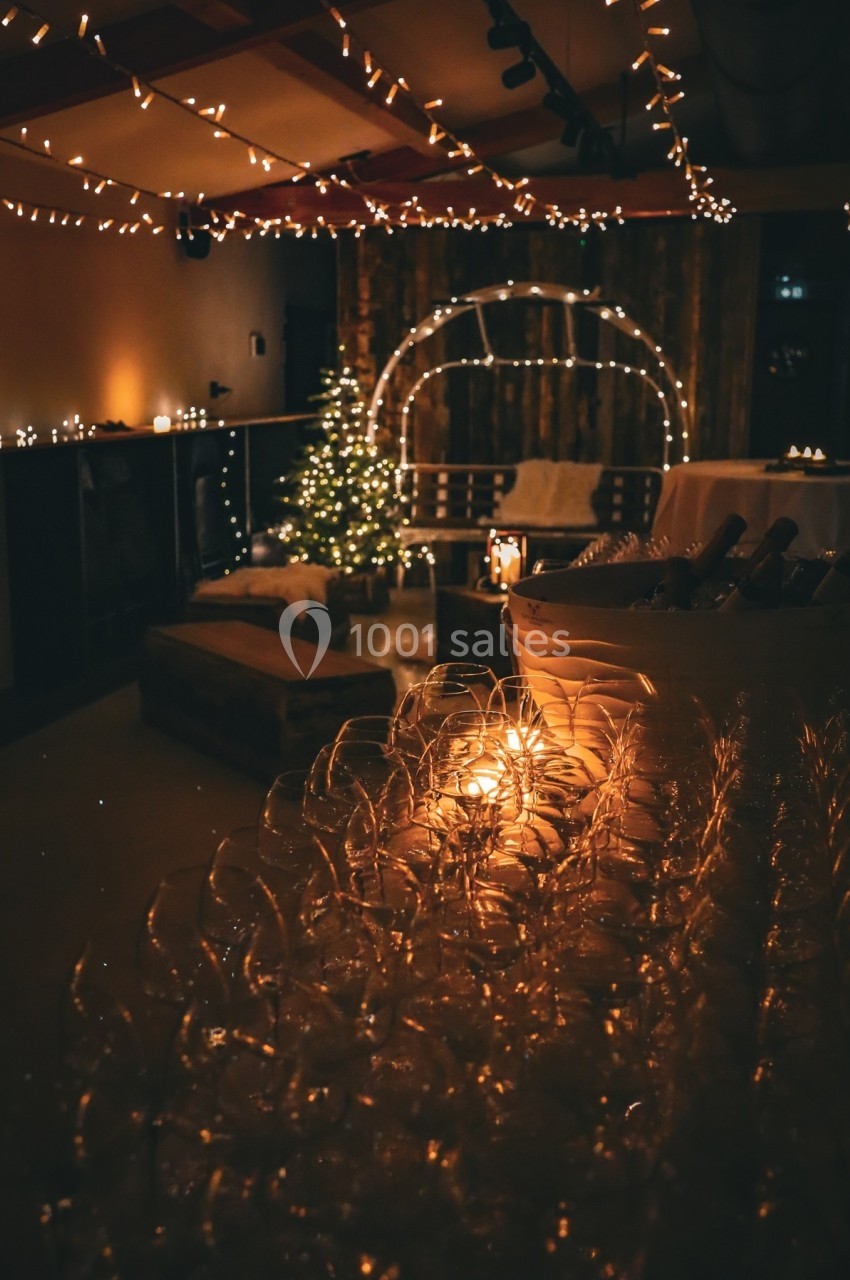 Salle décorée pour une fête avec guirlandes lumineuses, sapin de Noël et rangée de verres à vin au premier plan.