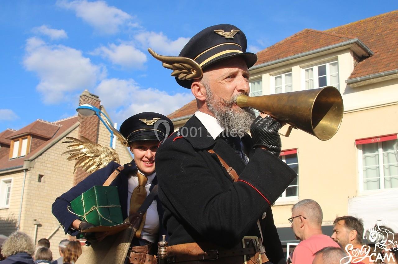 Un homme déguisé en aviateur joue du cornet en extérieur, accompagné d'une femme avec des ailes dorées.