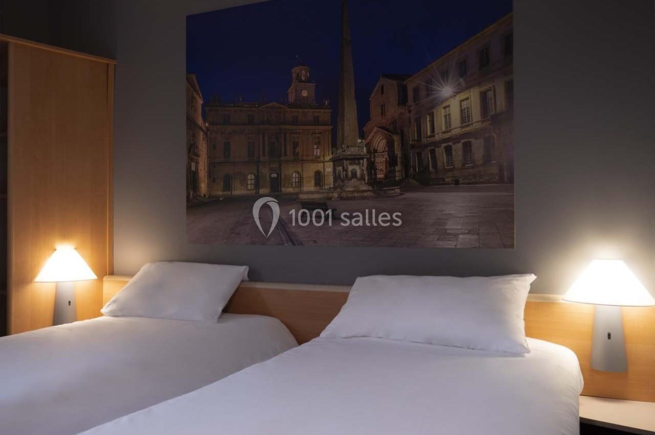 Deux lits simples avec une photo murale d'une place illuminée de nuit, dans une chambre sobrement éclairée.