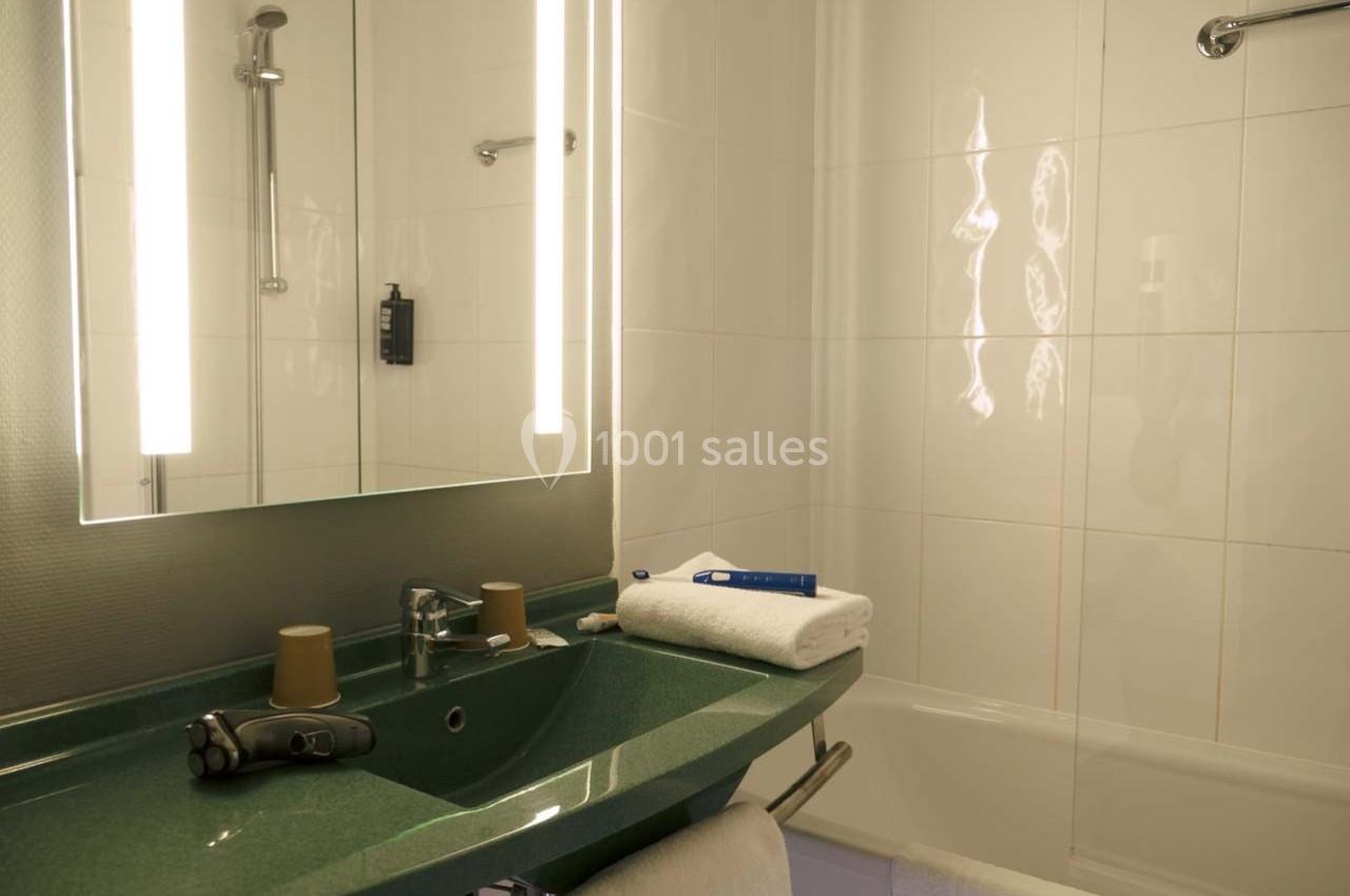 Salle de bain avec lavabo vert, miroir éclairé, serviette pliée, rasoir électrique et baignoire en arrière-plan.