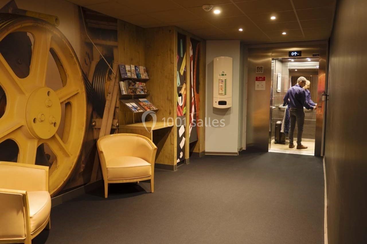 Homme entrant dans un ascenseur dans un couloir d'hôtel avec fauteuils, présentoir de brochures et décor mural industriel.
