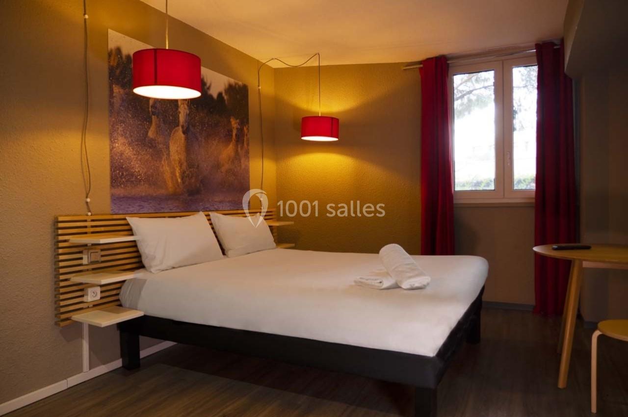 Chambre avec un lit double, draps blancs, deux lampes suspendues rouges et une fenêtre avec rideaux rouges.