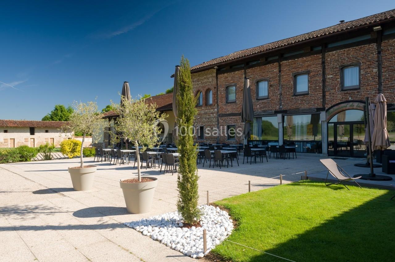 Terrasse extérieure d'un bâtiment en briques avec tables, chaises, parasols et végétation environnante.