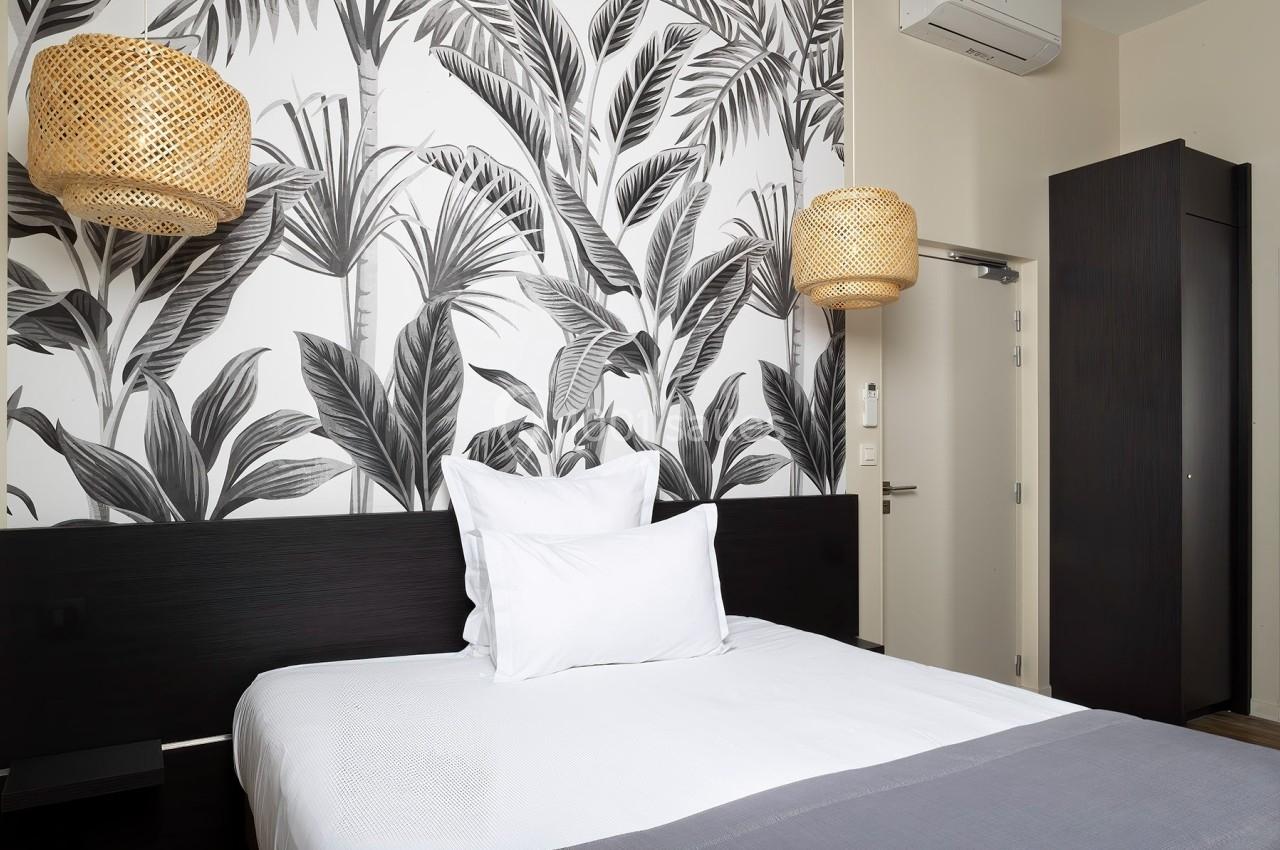 Lit double avec linge blanc, encadré par des lampes suspendues en rotin et un mur décoré de motifs végétaux noirs et blancs.