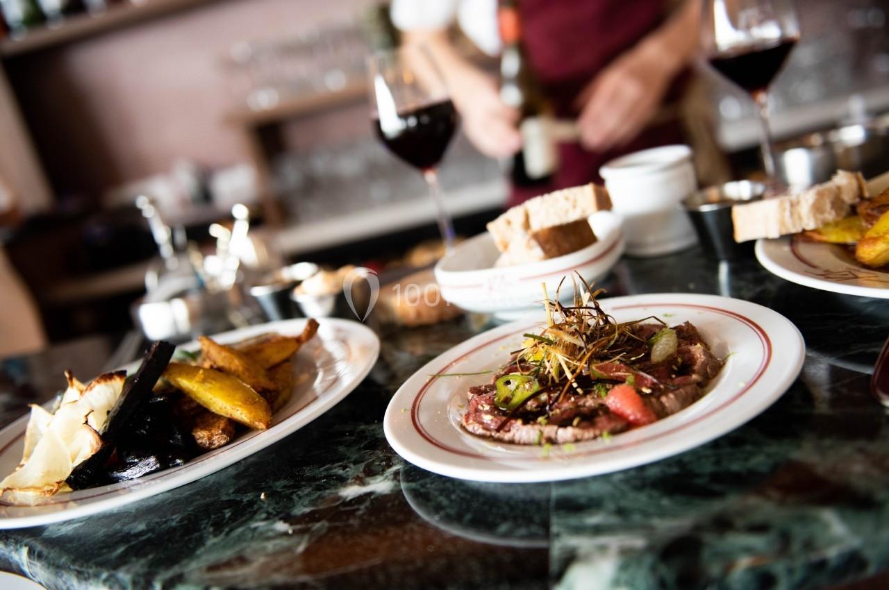 Assiette de viande garnie de légumes et d'herbes, entourée de plats d'accompagnement et de verres de vin sur une table.