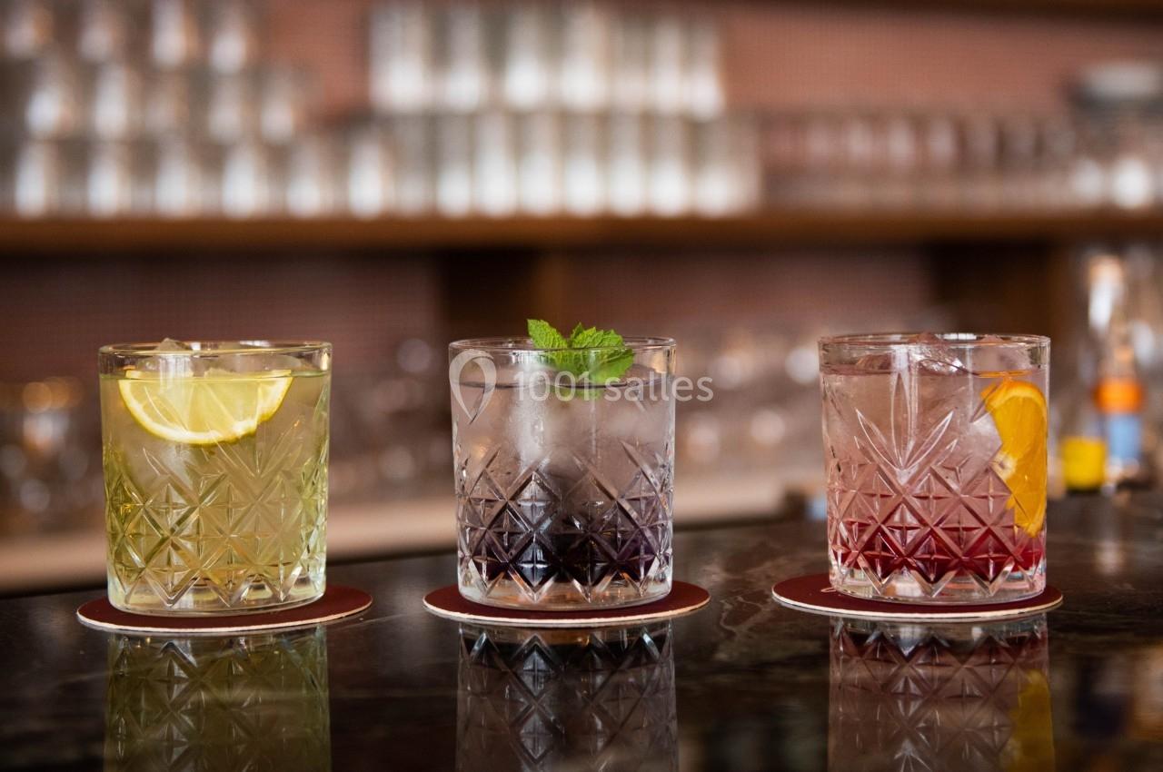 Trois verres de cocktails colorés garnis de tranches d'agrumes et de menthe, posés sur un comptoir de bar.