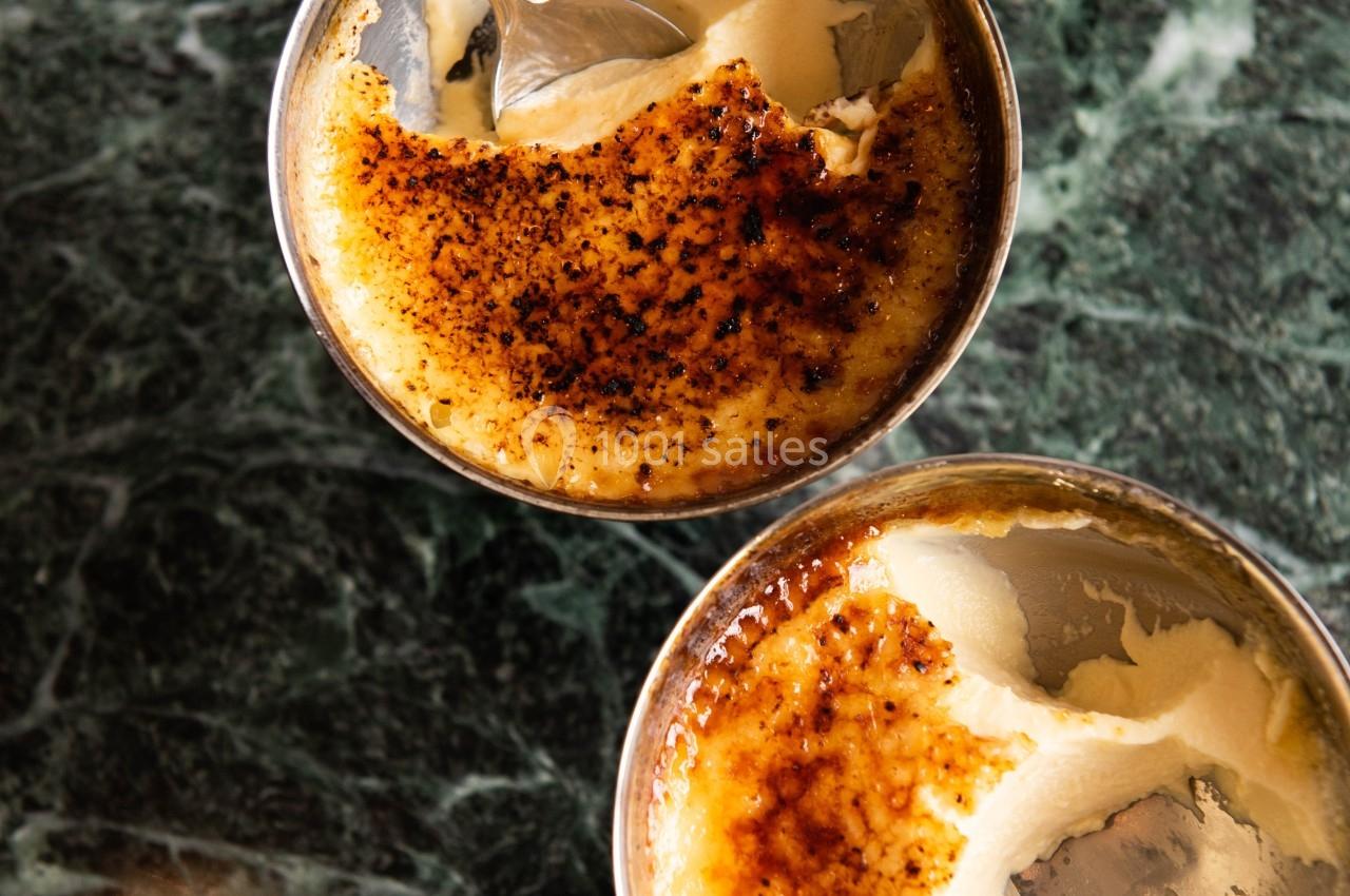 Deux crèmes brûlées avec une croûte caramélisée partiellement brisée, servies sur une table en marbre vert.