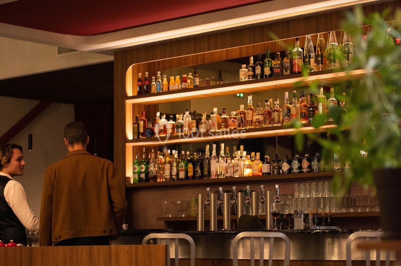 Un bar moderne avec des étagères remplies de bouteilles, deux personnes discutent au comptoir.