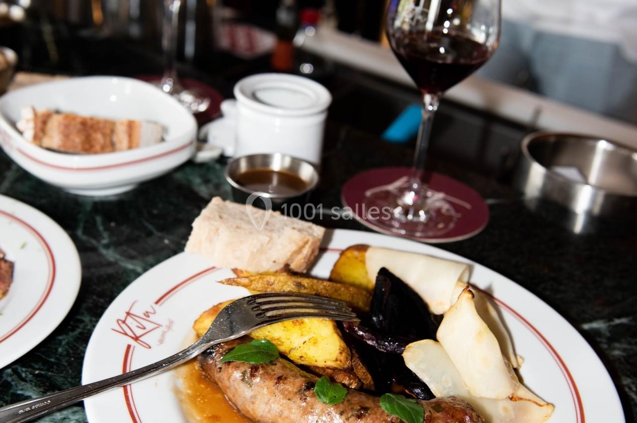 Assiette de saucisse avec légumes rôtis, sauce et pain, servie sur une table en marbre avec des verres de vin rouge.
