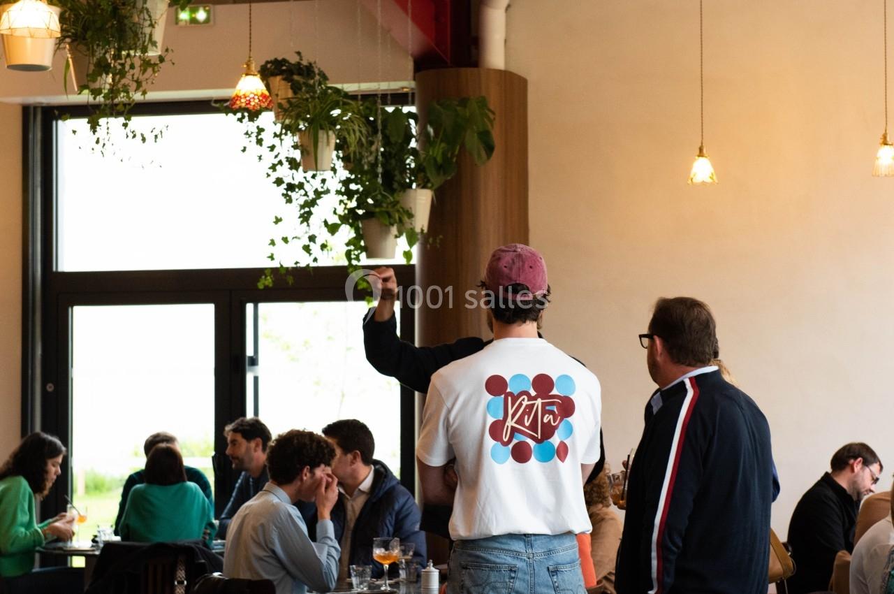 Des personnes discutent et prennent un verre dans un café lumineux avec des plantes suspendues et une grande baie vitrée.