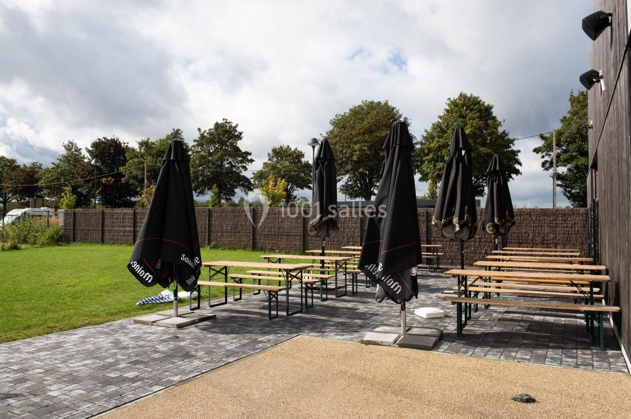 Terrasse extérieure avec tables en bois, bancs et parasols noirs, entourée d'une pelouse et de haies sous un ciel nuageux.