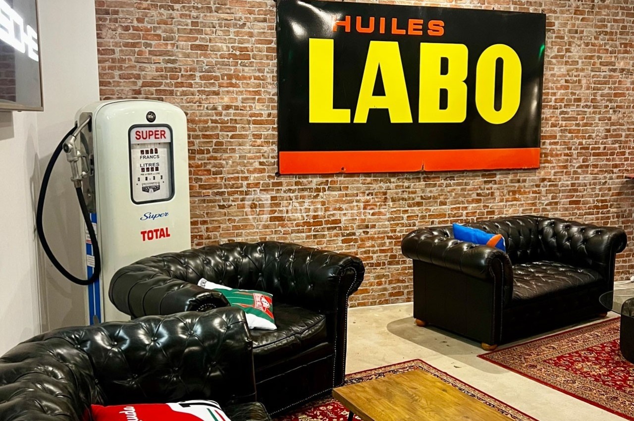 Salon rétro avec canapés en cuir, pompe à essence vintage et panneau mural ’Huiles Labo’ sur fond de briques.
