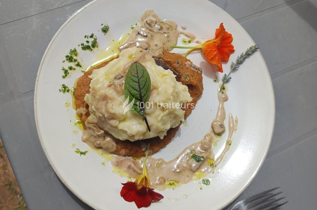 Assiette contenant un plat garni de purée, sauce crémeuse, fleurs comestibles et herbes fraîches sur une table grise.
