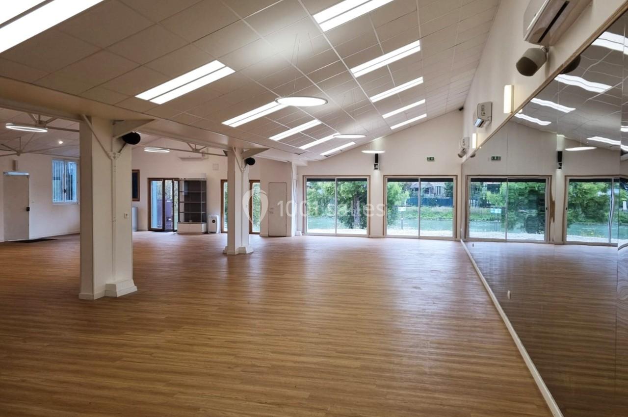 Salle spacieuse avec parquet clair, murs blancs, grandes baies vitrées et miroirs reflétant l'intérieur lumineux.