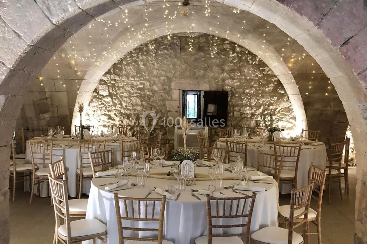 Salle voûtée en pierre décorée de guirlandes lumineuses, avec des tables rondes dressées pour un événement.