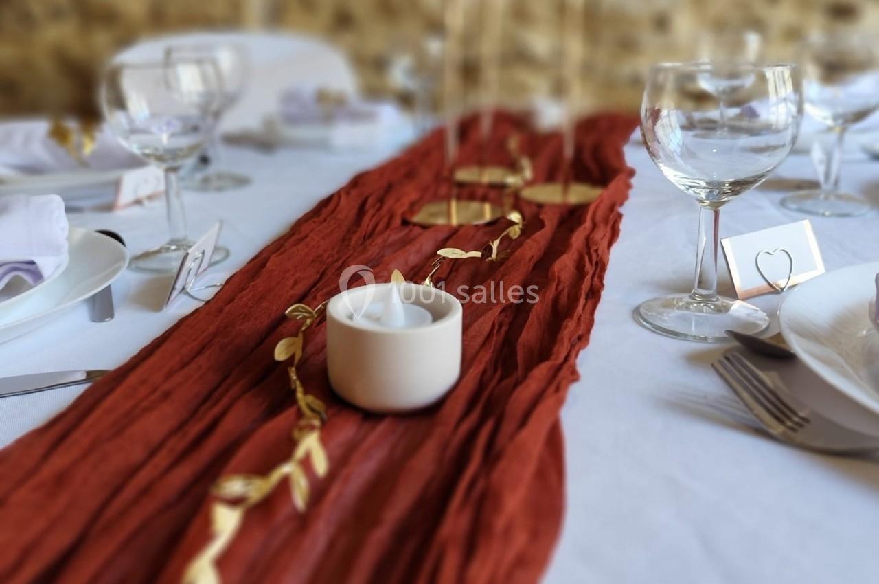 Chemin de table rouge texturé avec décoration dorée et bougie blanche, entouré de verres et assiettes sur une table dressée.