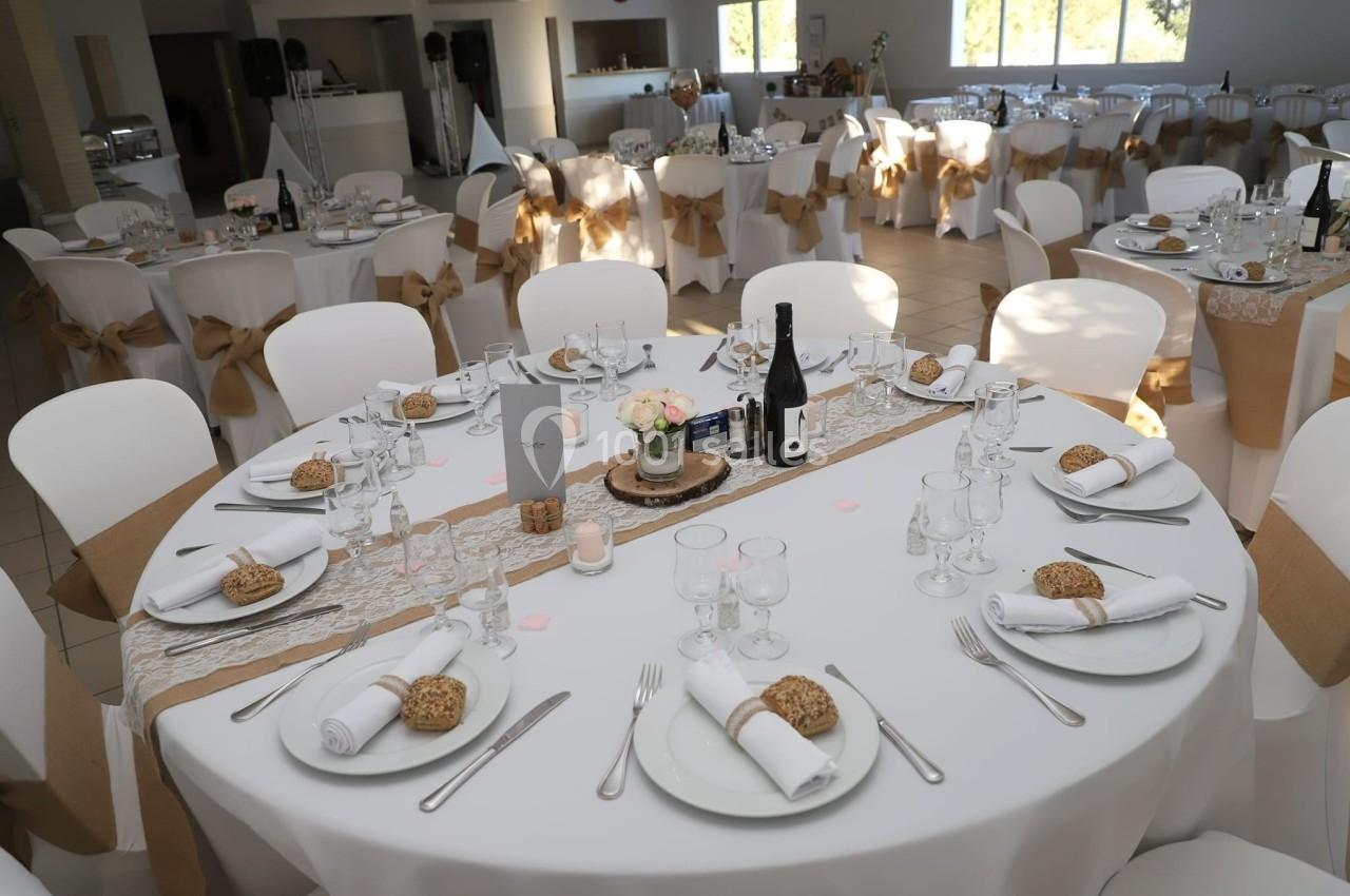 Salle de réception décorée avec des tables rondes dressées, nappes blanches et chaises ornées de nœuds beige.
