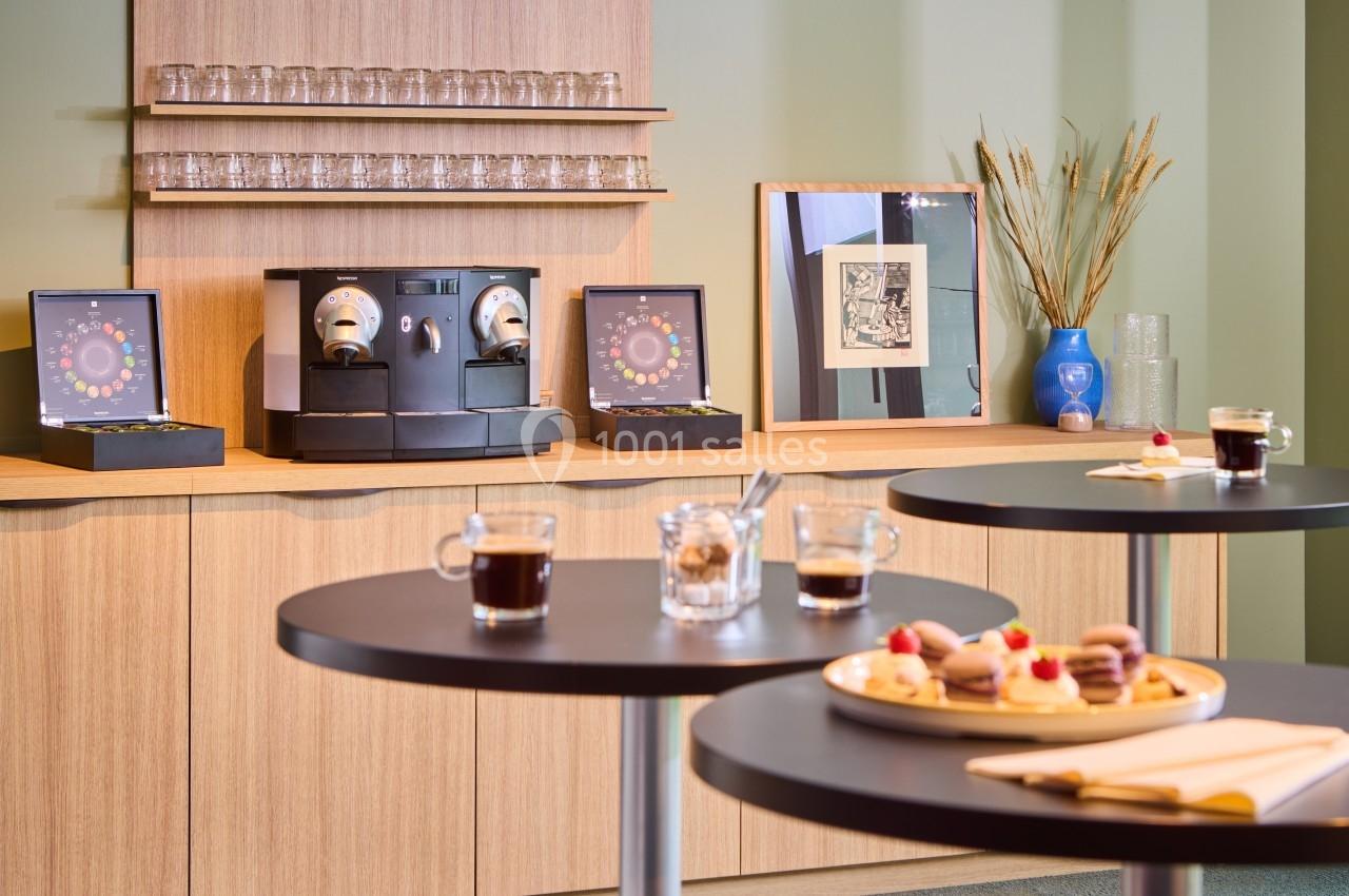 Machine à café avec tasses et capsules sur un comptoir en bois, tables avec desserts et boissons au premier plan.