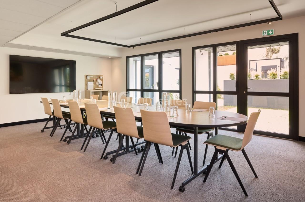 Salle de réunion lumineuse avec une grande table ovale, des chaises en bois et un écran mural.