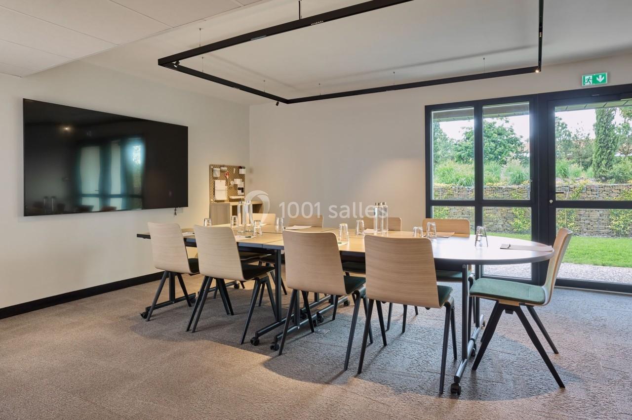 Salle de réunion lumineuse avec table rectangulaire, chaises modernes, écran mural et vue sur un jardin par une baie vitrée.