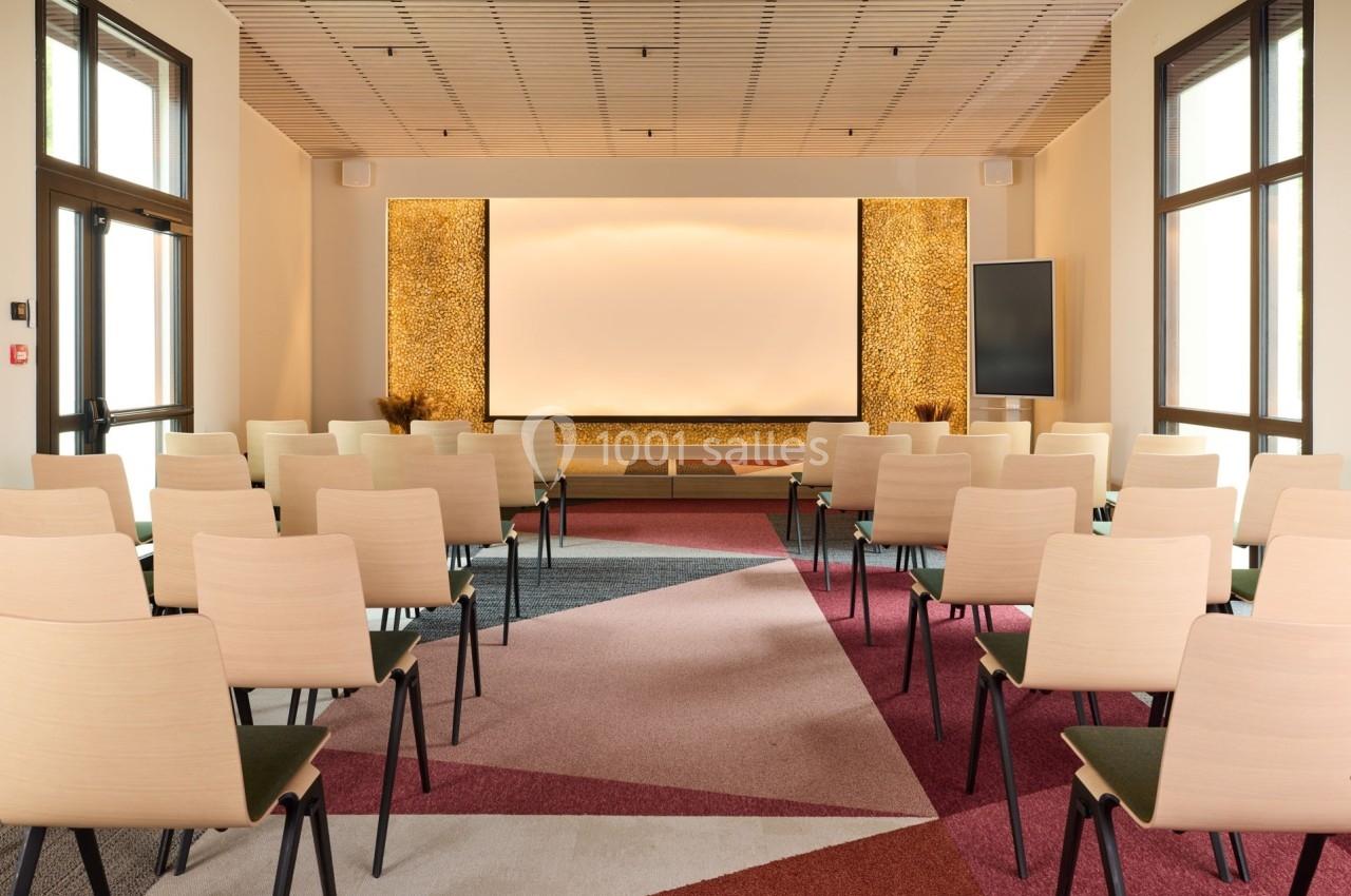 Salle de conférence lumineuse avec des rangées de chaises alignées face à un écran blanc et des murs décoratifs.