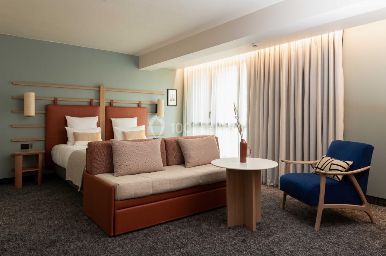 Chambre d'hôtel moderne avec lit double, canapé, fauteuil bleu, table ronde et grande fenêtre avec rideaux.
