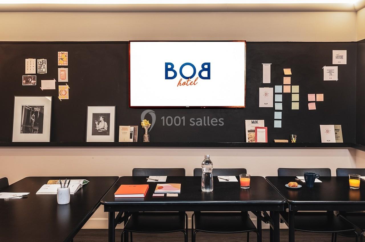 Salle de réunion avec tables noires, écran affichant ’BOB hôtel’ et mur décoré de cadres et notes colorées.