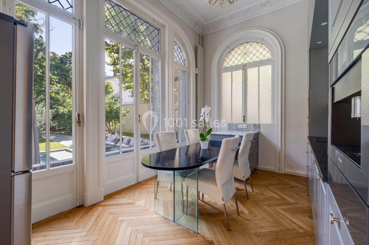 Salle à manger lumineuse avec table en verre, chaises blanches et grandes fenêtres donnant sur un jardin verdoyant.