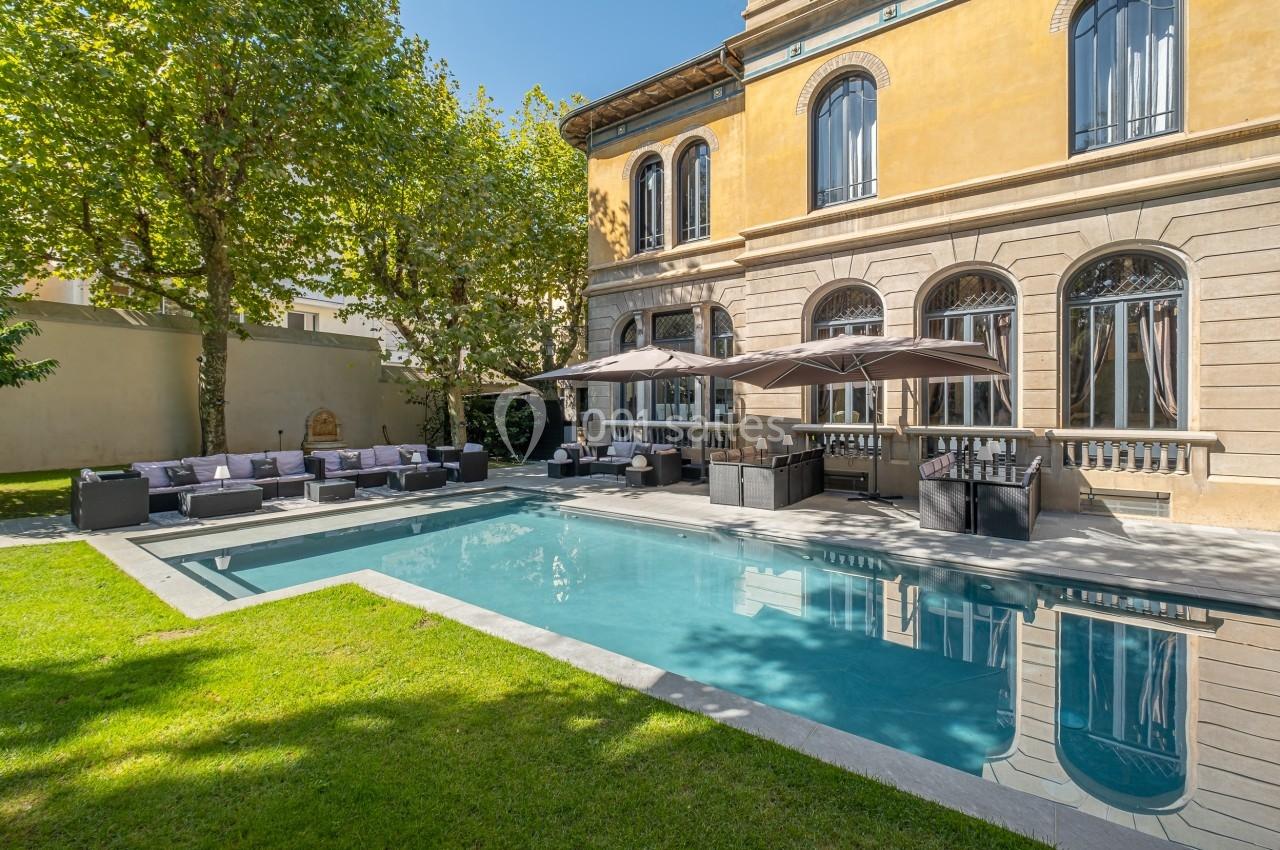 Piscine extérieure entourée de chaises longues, parasols et bâtiment ancien avec grandes fenêtres en arrière-plan.