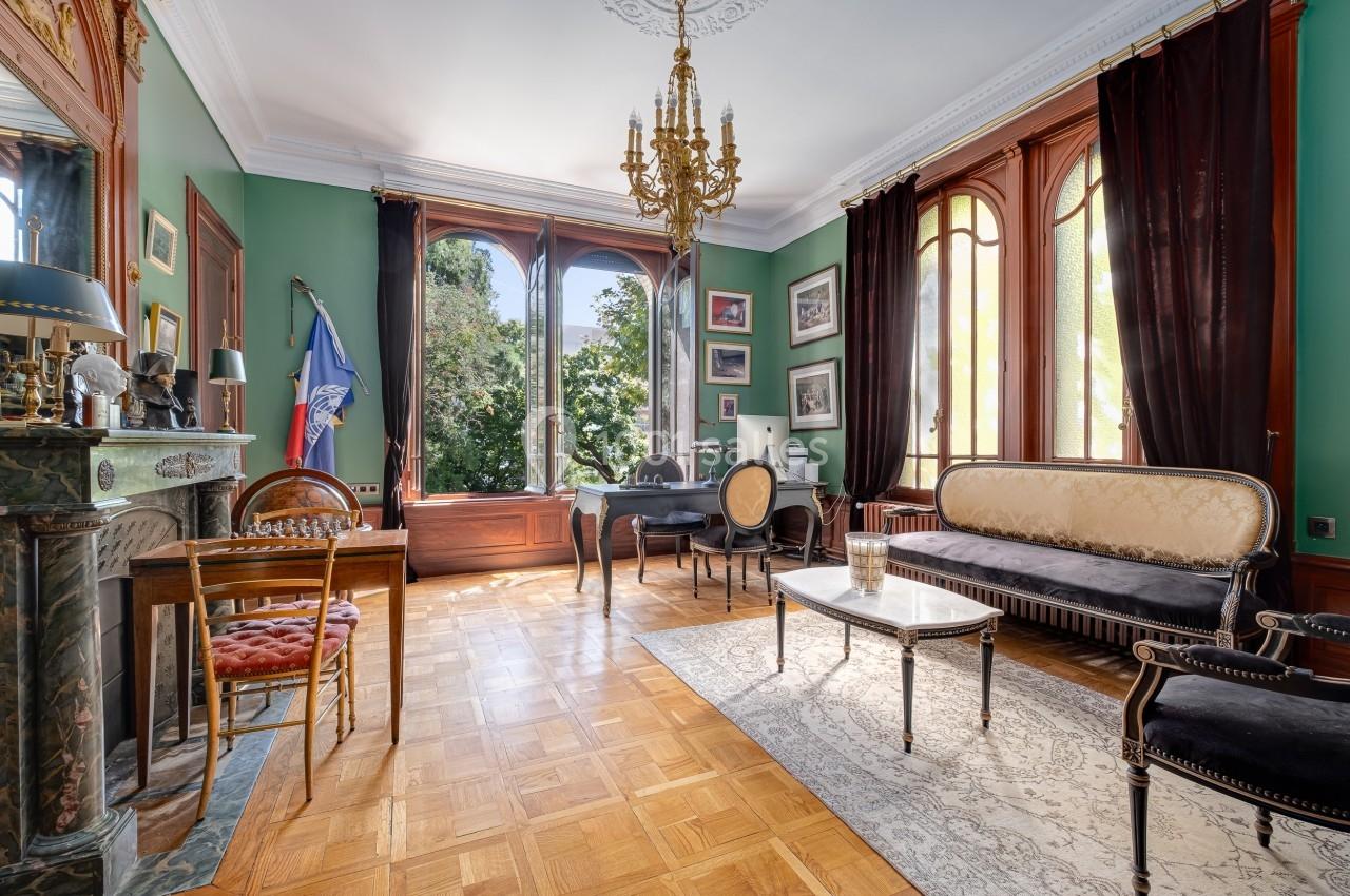 Salon élégant avec parquet, murs verts, grandes fenêtres, mobilier classique et cheminée ornée.