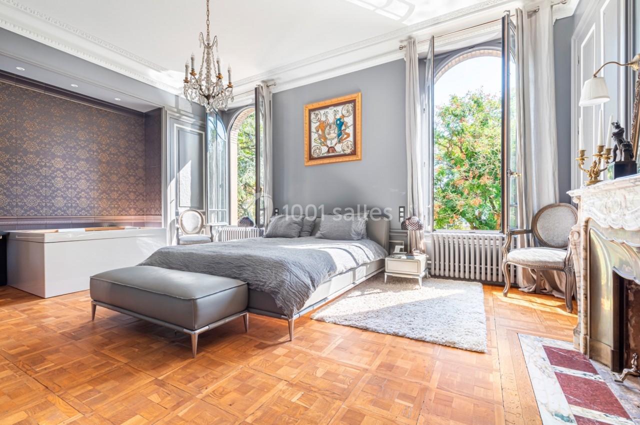 Chambre lumineuse avec lit double, parquet, cheminée, lustre, et grandes fenêtres donnant sur un espace arboré.