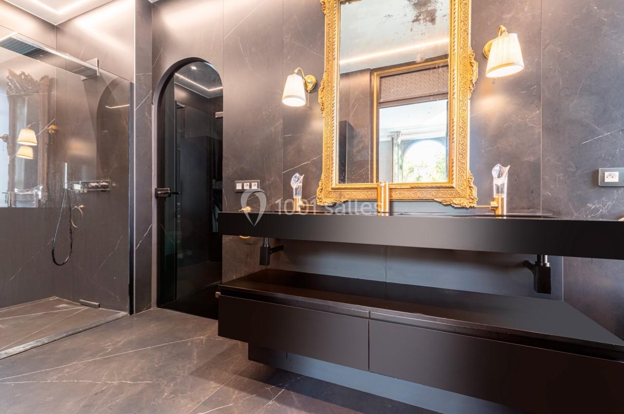 Salle de bain moderne avec murs sombres, miroir doré, double vasque noire et douche vitrée à gauche.