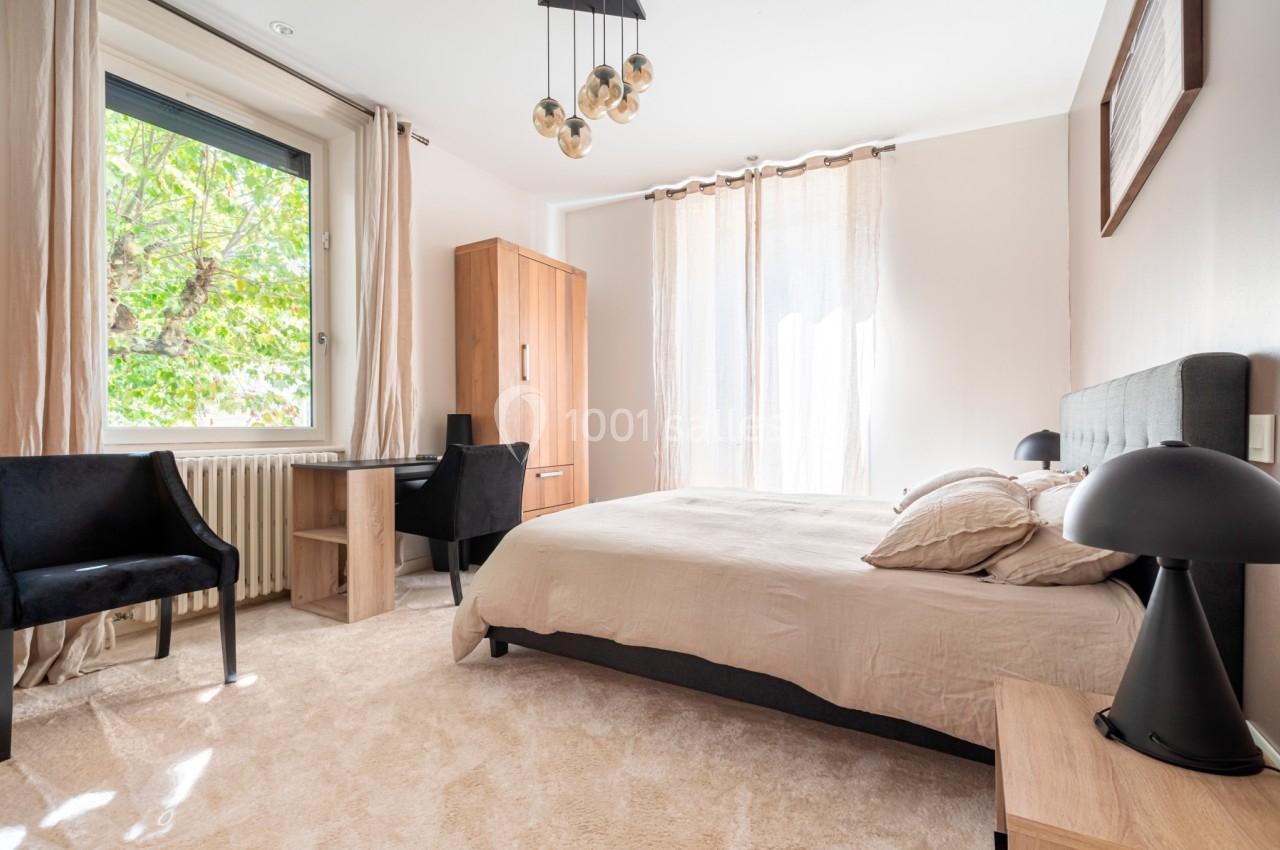 Chambre lumineuse avec lit double, bureau, deux chaises noires et grandes fenêtres donnant sur un arbre.