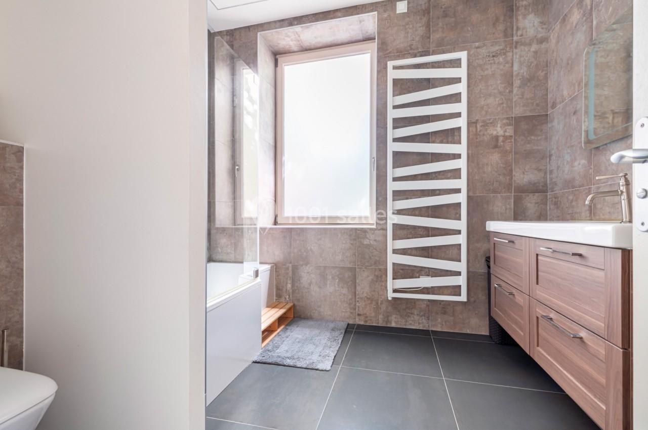 Salle de bain moderne avec carrelage marron, baignoire, meuble en bois clair et radiateur mural blanc.