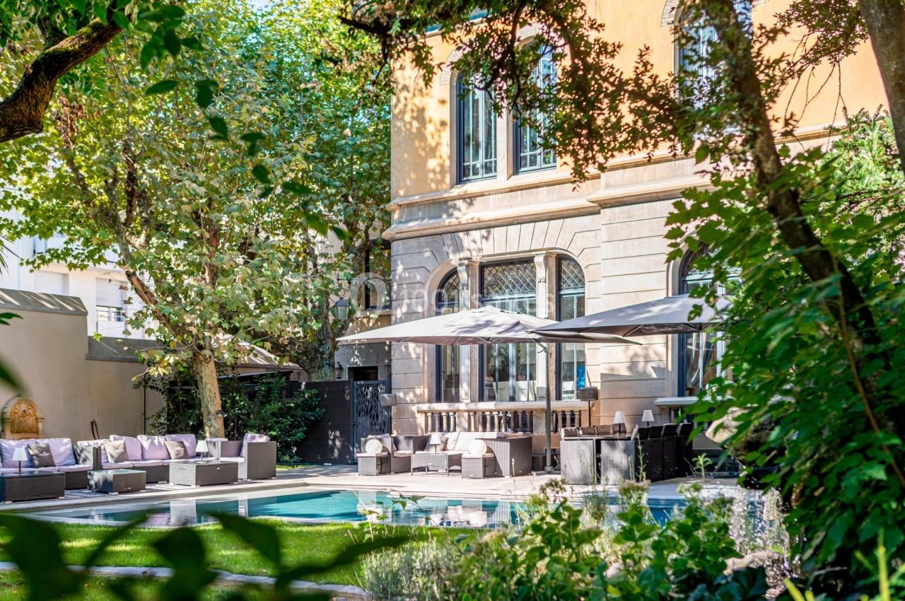 Jardin avec piscine, mobilier de salon extérieur et parasols, entouré de verdure devant un bâtiment aux grandes fenêtres.