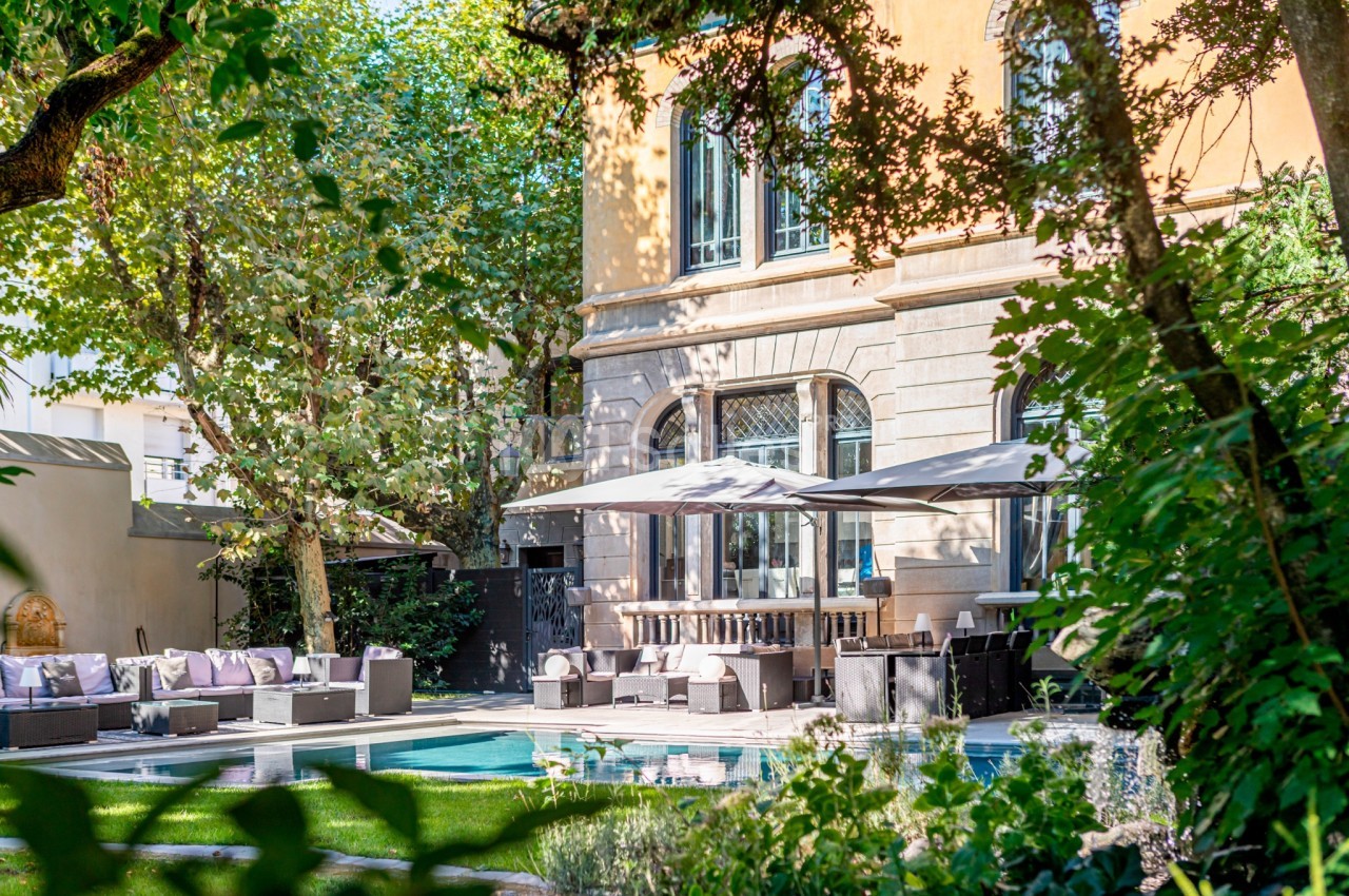 Jardin avec piscine, mobilier de salon extérieur et parasols, entouré de verdure devant un bâtiment aux grandes fenêtres.