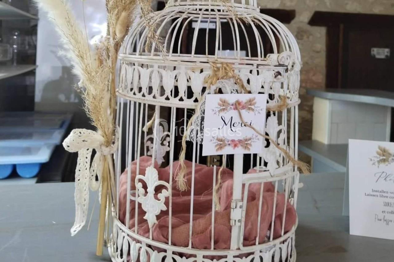 Cage décorative blanche avec fleurs séchées, rubans et coussin rose, posée sur une table grise.