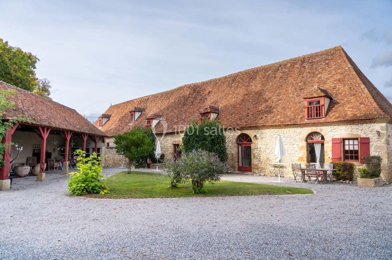 Bâtisse en pierre avec toit en tuiles, entourée de végétation et d'une cour gravillonnée, sous un ciel légèrement nuageux.