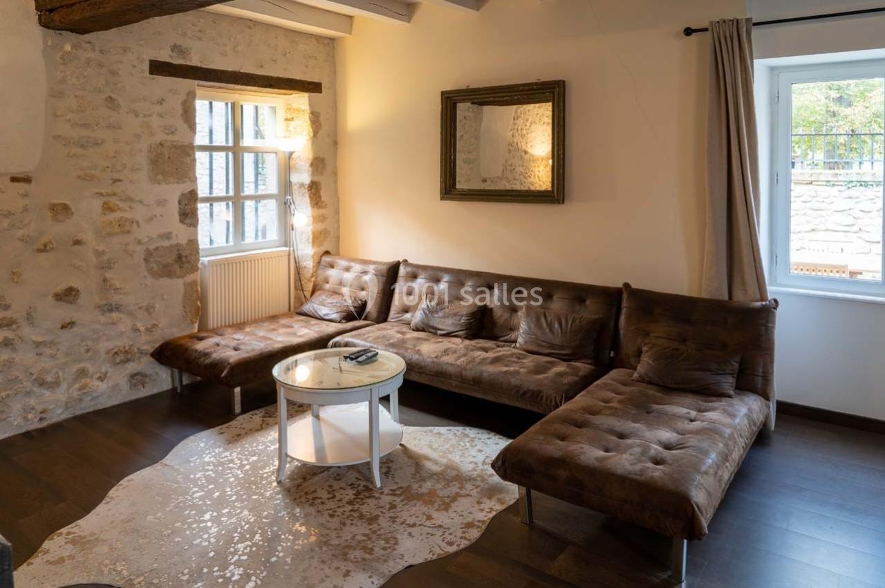 Salon avec murs en pierre, canapé en cuir marron en angle, table basse ronde et lumière naturelle entrant par les fenêtres.