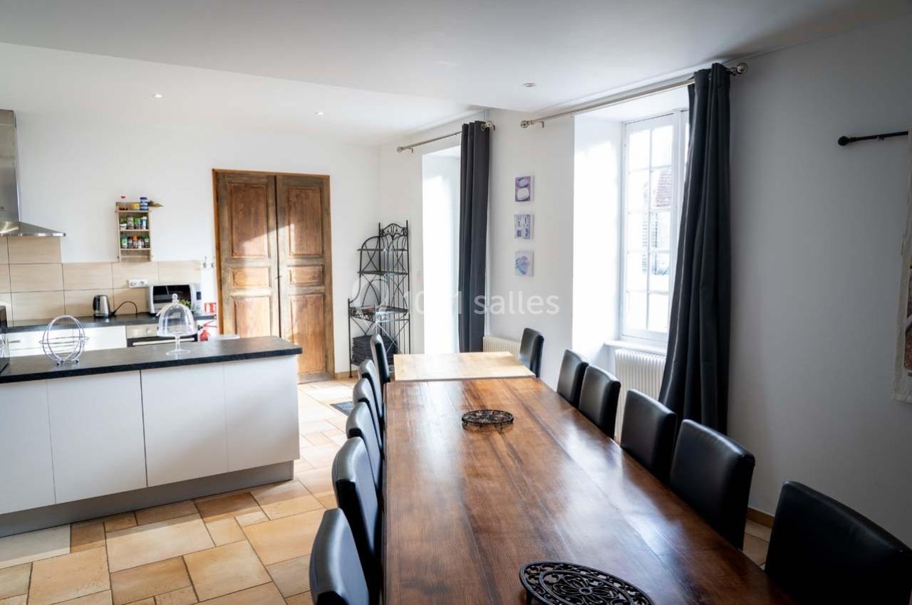 Cuisine lumineuse avec une grande table en bois, des chaises noires, un îlot central et des portes en bois au fond.