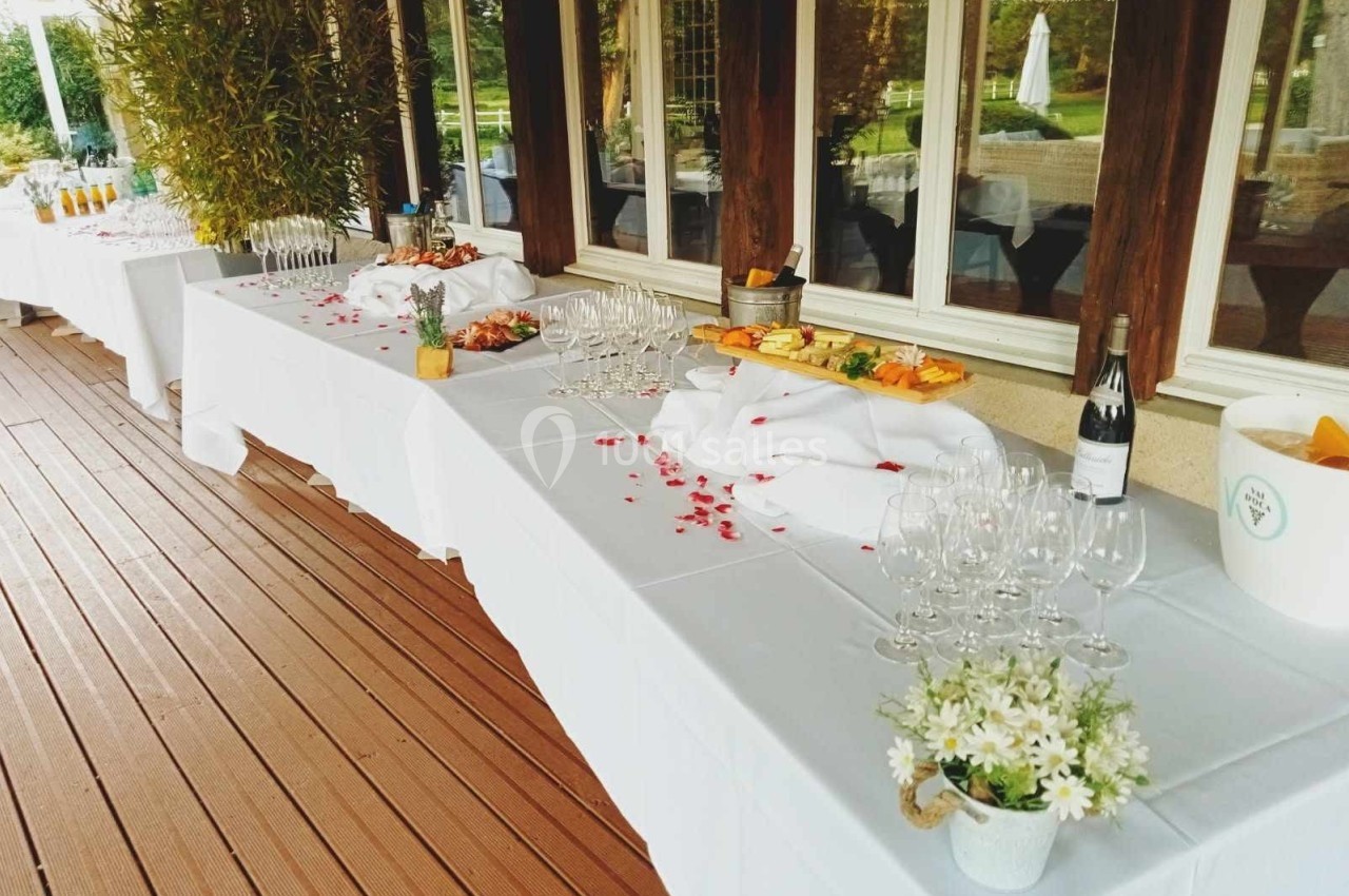 Tables dressées avec nappes blanches, verres, bouteilles et plats variés sur une terrasse en bois près de grandes fenêtres.