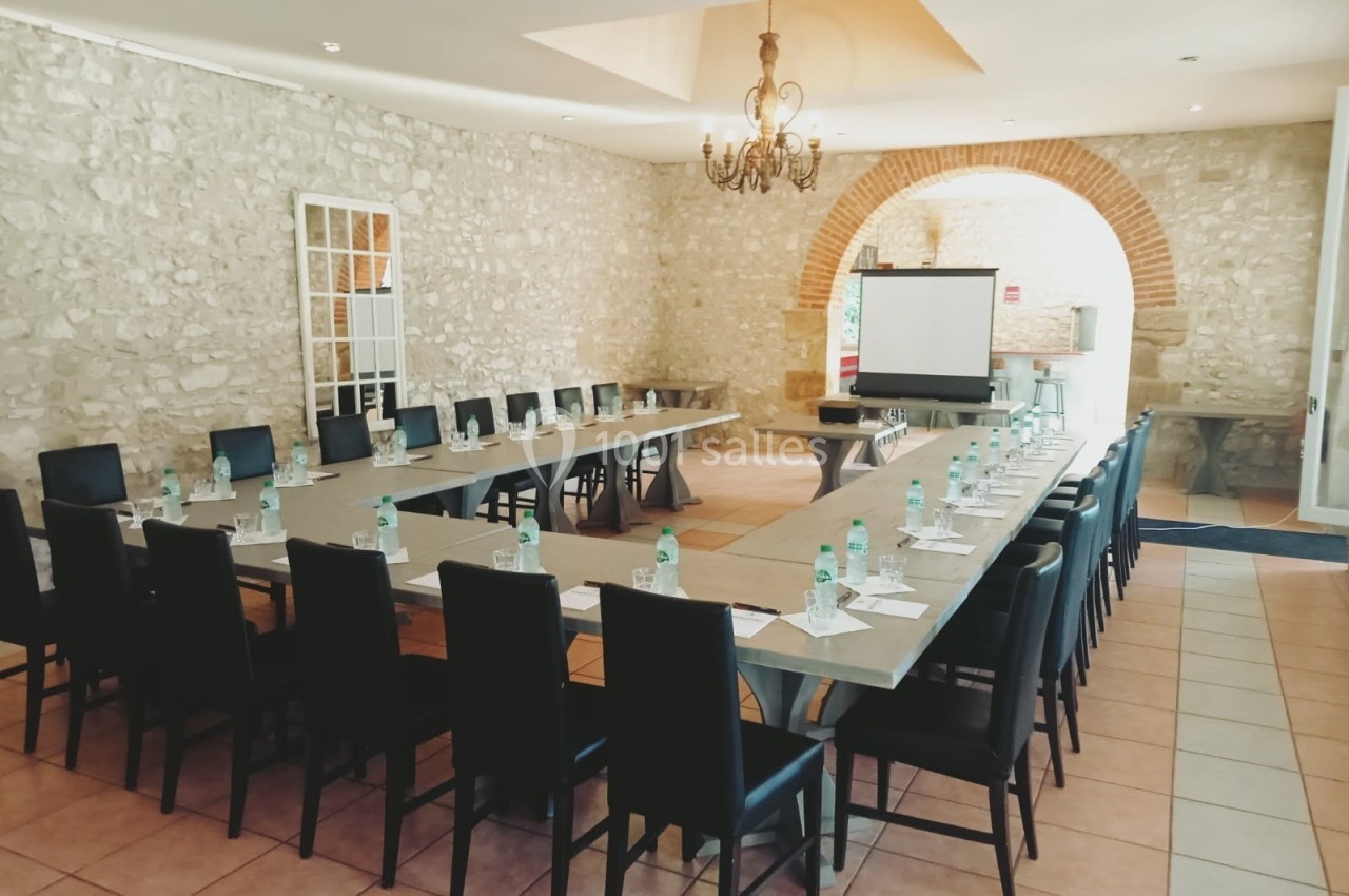 Salle de réunion lumineuse avec murs en pierre, tables disposées en U, chaises noires et écran de projection.