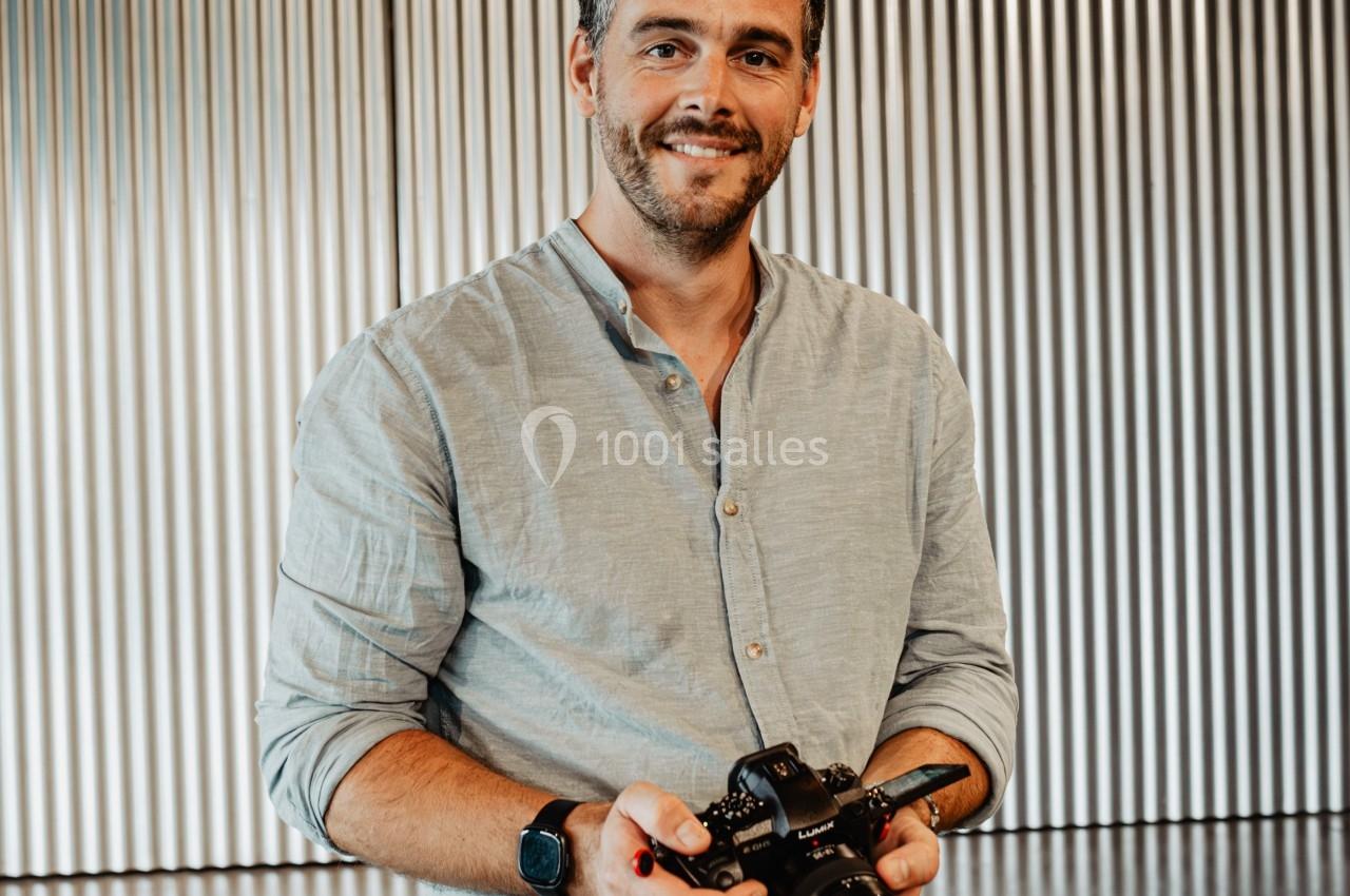 Homme souriant tenant un appareil photo dans un espace intérieur avec un fond de rideaux métalliques.