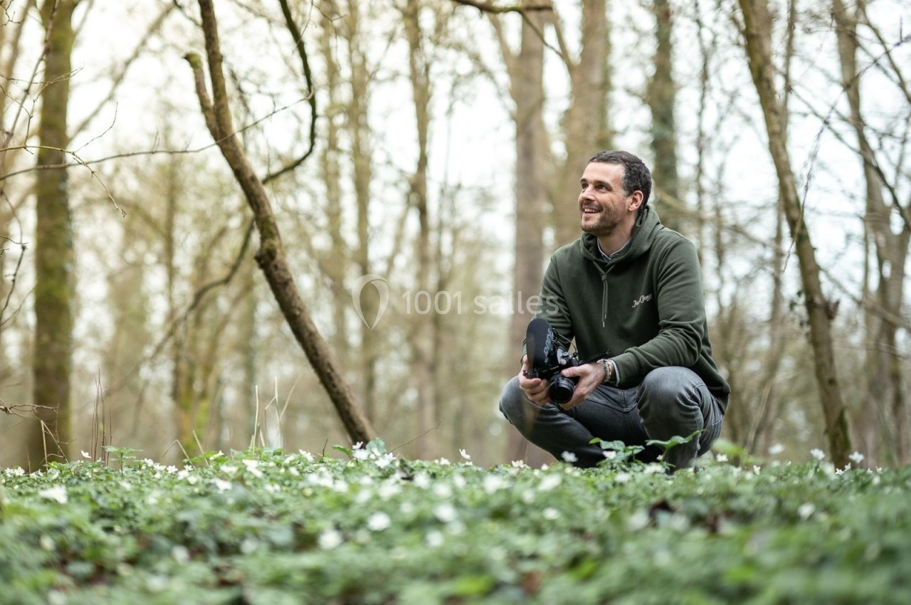 Un homme accroupi dans une forêt, tenant un appareil photo, entouré de végétation et de petites fleurs blanches.